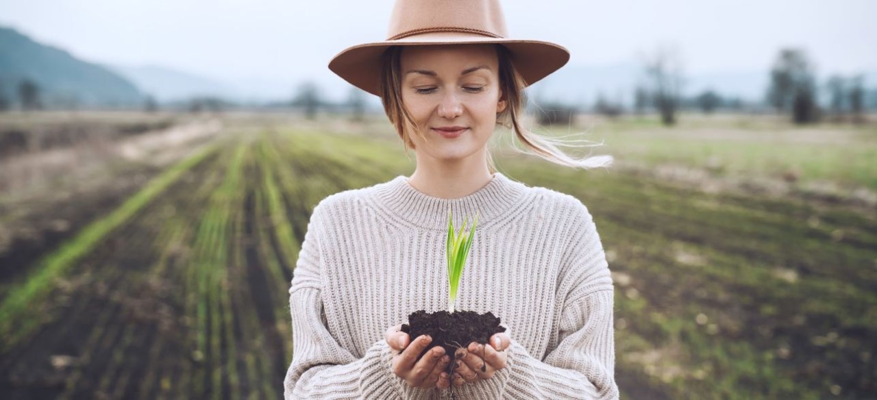 Mujer joven en un cultivo, sosteniendo en sus manos una planta mientras piensa cómo hacer un planeta sostenible.